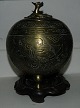 Skål i metal (bronce ?) på tre fødder. Stående på base af træ. Dekoreret med mænd til hest på ...